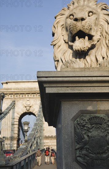 Budapest, Pest County, Hungary. Lion statue on Szechenyi Chain Bridge or Memory Bridge with passing pedestrians seen behind. Hungary Hungarian Europe European East Eastern Buda Pest Budapest City Architecture Transport Bridge Szechenyi Chain Memory Walking people Pedestrians Tourists Statue Lion Stone Destination Destinations Eastern Europe Holidaymakers Tourism