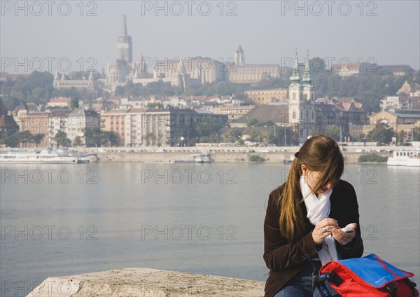Budapest, Pest County, Hungary. Young woman using mobile phone on Pest bank of the River Danube with Buda Castle behind. Hungary Hungarian Europe European East Eastern Buda Pest Budapest City Young Woman Female Girl Using Personal Organiser PDA Mobile Phone Cell Telephone Communications River Danube Water Castle Castillo Castello Cellular Eastern Europe Female Women Girl Lady Immature One individual Solo Lone Solitary