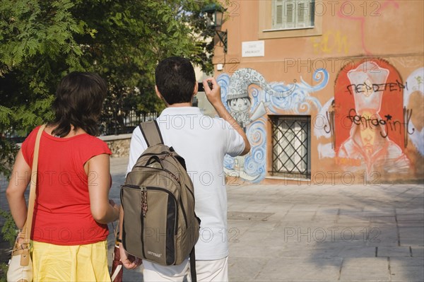 Athens, Attica, Greece. Tourist couple photographing graffiti on building in Plaka district lying just beneath the Acropolis. Atenas Athenes Destination Destinations Ellada European Greek Holidaymakers Sightseeing Southern Europe Tourism Tourists