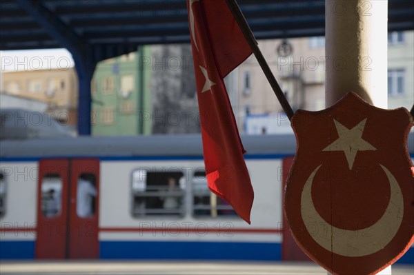 Istanbul, Turkey. Sultanahmet. Istanbul Sirkeci Terminal Turkish flag and emblem in foreground with train on platform behind. Sirkeci GarA is a terminus main station of the Turkish State Railways or TCDD in Sirkeci on the European part of Istanbul. International domestic and regional trains running westwards depart from this station which was inaugurated as the terminus of the Orient Express. Turkey Turkish Istanbul Constantinople Stamboul Stambul City Europe European Asia Asian East West Urban Destination Travel Tourism Transportt Rail Railway Train Trains Station Platform Flag Emblem Commuter Sirkeci TCCD Color Destination Destinations Middle East South Eastern Europe Turkiye Western Asia