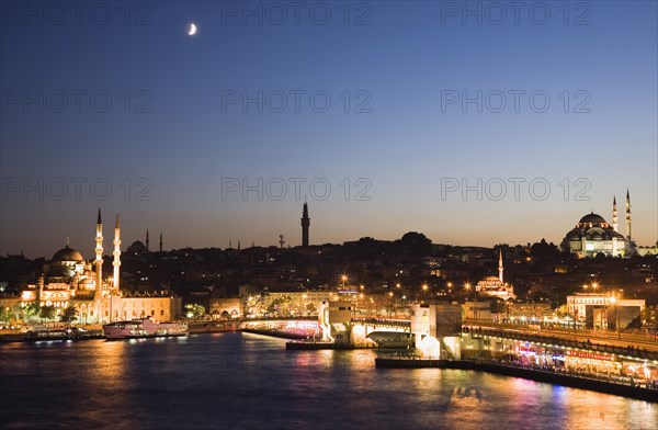 Istanbul, Turkey. Sultanahmet. The Golden Horn. The New Mosque or Yeni Camii at left the Galata Bridge and Suleymaniye Mosque at right illuminated at dusk with crescent moon in sky above and coloured lights reflected in the water. Turkey Turkish Istanbul Constantinople Stamboul Stambul City Europe European Asia Asian East West Urban Skyline Destination Travel Tourism Sultanahmet Golden Horn New Mosque Yeni Camii Galata Bridge Suleymaniye Dusk Sunset Night Illuminated Skyline Minarets Water Blue Citiscape Building Buildings Urban Architecture Colored Destination Destinations Middle East Nightfall Twilight Evenfall Crespuscle Crespuscule Gloam Gloaming Nite South Eastern Europe Sundown Atmospheric Turkiye Western Asia