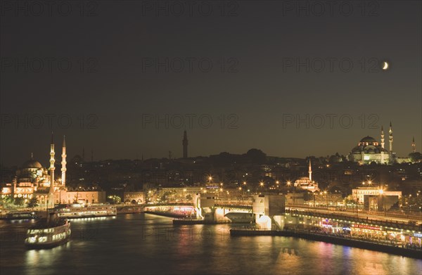 Istanbul, Turkey. Sultanahmet. The Golden Horn. The New Mosque or Yeni Camii at left the Galata Bridge and Suleymaniye Mosque at right illuminated at night with crescent moon in sky above. Turkey Turkish Istanbul Constantinople Stamboul Stambul City Europe European Asia Asian East West Urban Skyline Destination Travel Tourism Sultanahmet Golden Horn New Mosque Yeni Camii Galata Bridge Suleymaniye Dusk Sunset Night Illuminated Skyline Minarets Water Crescent Moon Blue Citiscape Building Buildings Urban Architecture Destination Destinations Middle East Nightfall Twilight Evenfall Crespuscle Crespuscule Gloam Gloaming Nite South Eastern Europe Sundown Atmospheric Turkiye Western Asia