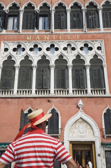 Venice, Veneto, Italy. Cropped shot of gondolier wearing red and white striped shirt and straw boater trimmed with red sash standing in front of facade of the Hotel Danieli. Italy Italia Italian Venice Veneto Venezia Europe European City Red Gondolier Man Striped Stripe Stripes Architecture Facade Hotel Danieli Hat Sash Ribbon Band Window Windows Shutters Color Destination Destinations Male Men Guy One individual Solo Lone Solitary Southern Europe