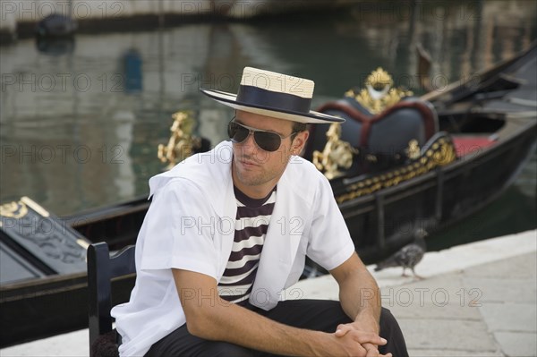 Venice, Veneto, Italy. Centro Storico Young gondolier in traditional uniform of striped shirt and straw hat with black sash wearing sunglasses seated beside moored gondola on canal in late summer sunshine. Italy Italia Italian Venice Veneto Venezia Europe European City Sun Shades Sunglasses Canal Water Boater Hat Man People Gondolier Gondolas Gondola Storico Centro Classic Classical Destination Destinations Historical Immature Male Men Guy Older One individual Solo Lone Solitary Southern Europe