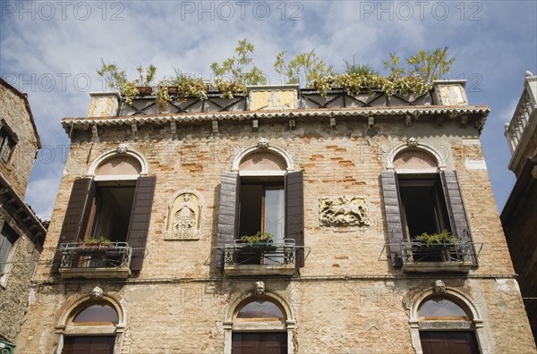 Venice, Veneto, Italy. Building facade with multiple windows each with small balcony window box and wooden shutters. Plaque depicting St. George and the Dragon and pots of plants on rooftop above. Italy Italia Italian Venice Veneto Venezia Europe European City Architecture Facade Exterior Windows Shutters Flowers Destination Destinations Southern Europe