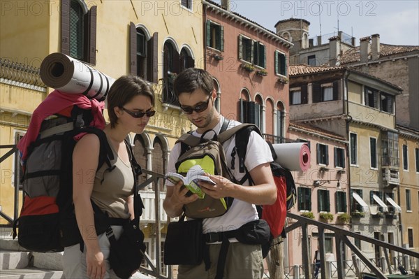 Venice, Veneto, Italy. Two young backpacker tourists wearing sunglasses and carrying rucksacks rolled up sleeping mats and moneybelts consulting guide book while standing on canal bridge in Centro Storico centre of Venice. Italy Italia Italian Venice Veneto Venezia Europe European City Tourist Tourists Bridge Architecture Map Guide Book Backpackers Rucksacks Knapsacks 2 Center Destination Destinations Holidaymakers Immature Sightseeing Southern Europe Tourism