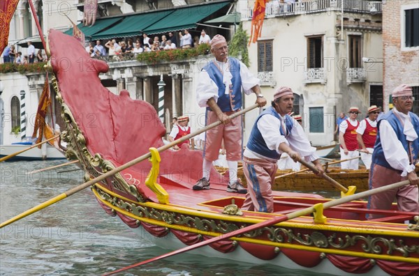 Venice, Veneto, Italy. Participants in the Regatta Storico historical annual regatta wearing traditional costume rowing gondola past onlookers gathered on the balconies of canalside buildings behind. Italy Italia Italian Venice Veneto Venezia Europe European City Regata Regatta Gondola Gondola Gondolas Gondolier Boat Architecture Exterior Water Classic Classical Destination Destinations History Historic Holidaymakers Older Southern Europe Tourism Tourist