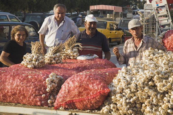 Kusadasi, Aydin Province, Turkey. Stallholder selling garlic at weekly market standing behind stall piled with sacks of garlic bulbs in late afternoon summer sunshine. Turkey Turkish Eurasia Eurasian Europe Asia Turkiye Aydin Province Kusadasi Market markets Stall Vendor Man Male Men Garlic Sacks Bulb Bulbs Destination Destinations European Male Man Guy Male Men Guy Middle East South Eastern Europe Western Asia