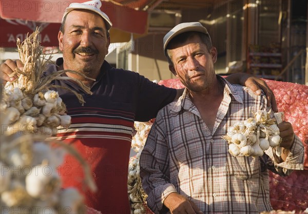 Kusadasi, Aydin Province, Turkey. Stallholders at weekly market with sacks of garlic for sale and holding up bunches of bulbs in late afternoon summer sunshine. Turkey Turkish Eurasia Eurasian Europe Asia Turkiye Aydin Province Kusadasi Market markets Stall Vendor Man Male Men Garlic Sacks Bulb Bulbs Destination Destinations European Male Man Guy Male Men Guy Middle East South Eastern Europe Western Asia