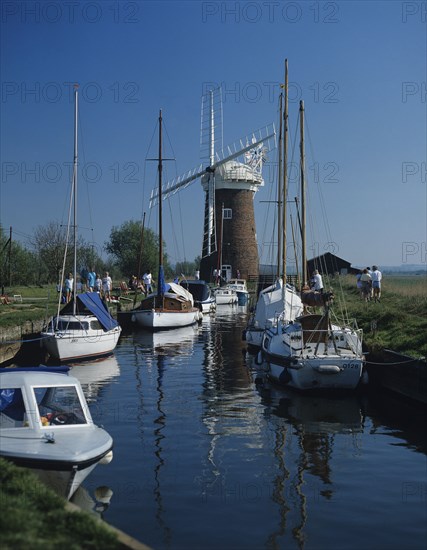 The Norfolk Broads, Norfolk, England. People walking along path next to canal with moored pleasure boats. England English UK United Kingdom GB Great Britain British Europe European Norfolk County Broads People Group Tourists Walk Walking Path Canal Water Boats Yachts Pleasure Leisure Windmill Blue Sky Waterway Waterways British Isles Destination Destinations Great Britain Holidaymakers Northern Europe Tourism United Kingdom