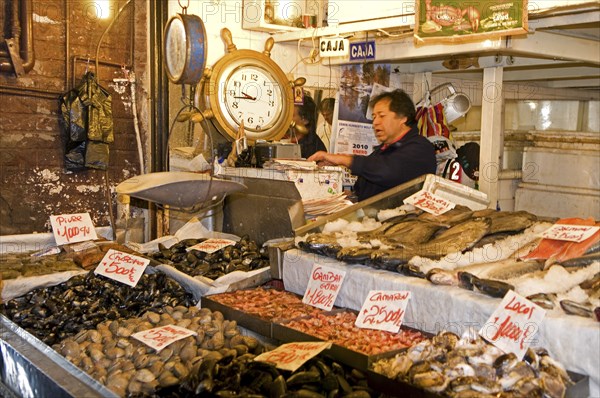 Santiago, Chile. Mecardo Central. Shellfish fresh fish and octopus on sale with prices clearly displayed. Vendor behind counter by a large clock and weighing scale. With a 5 million ton annual catch the country is the seventh-largest fishing nation in the world. Chile Chilean South America American Hispanic Santiago City Cityscape Urban Travel Destination Vacation Holiday Mercado Central Market Fish Seafood Shellfish Scales Clock Destination Destinations Five Latin America Latino One individual Solo Lone Solitary South America Southern