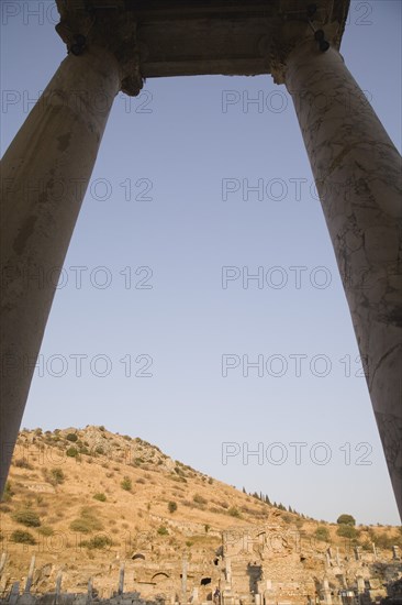 Selcuk, Izmir Province, Turkey. Ephesus. Pair of marble columns supporting roof framing ruins scattered across hillside in antique city of Ephesus on the Aegean sea coast. Turkey Turkish Eurasia Eurasian Europe Asia Turkiye Izmir Province Selcuk Ephesus Ruin Ruins Roman Facde Ancient Architecture Masonry Rock Stone Destination Destinations European History Historic Middle East Scenic South Eastern Europe Water Western Asia