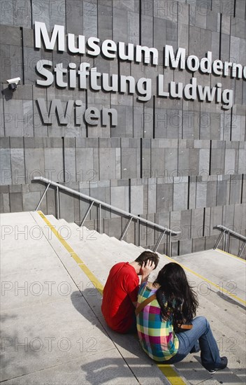 Vienna, Austria. Neubau District. Young student couple sitting on the steps of the Museum of Modern Art Museum Moderner Kunst Stiftung Ludwig Wien or MUMOK. Austria Austrian Republic Vienna Viennese Wien Europe European City Capital Neubau District Quarter Museums Mueseumsquartier Luwig Stiftung Kunst Moderner Museum Facade Exterior Architecture Mumok Couple Color Destination Destinations Gray Immature Osterreich Viena Western Europe