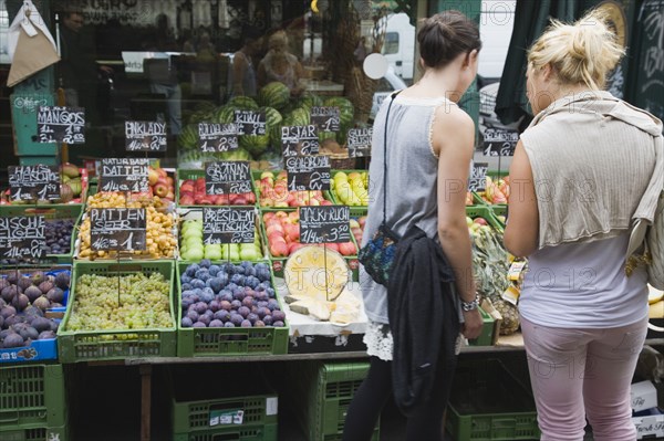 Vienna, Austria. The Naschmarkt. Two young women choosing fresh fruit from display including figs grapes and apples outside glass shopfront. Austria Austrian Republic Vienna Viennese Wien Europe European City Capital Naschmarkt Market Display Fresh Food Fruit Vegetables Veg Vegetable Stall Shop Store 2 Destination Destinations Female Woman Girl Lady Immature Osterreich Viena Western Europe