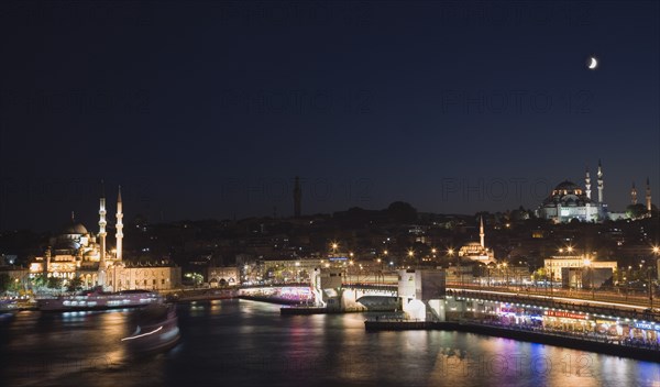 Istanbul, Turkey. Sultanahmet. The Golden Horn. The New Mosque or Yeni Camii at left the Galata Bridge and Suleymaniye Mosque at right illuminated at night with boats on the Bosphorus in motion blur and coloured lights reflected in the water. Turkey Turkish Istanbul Constantinople Stamboul Stambul City Europe European Asia Asian East West Urban Skyline Destination Travel Tourism Sultanahmet Golden Horn New Mosque Yeni Camii Galata Bridge Suleymaniye Dusk Sunset Night Illuminated Skyline Minarets Water Citiscape Building Buildings Urban Architecture Colored Destination Destinations Middle East Nightfall Twilight Evenfall Crespuscle Crespuscule Gloam Gloaming Nite South Eastern Europe Sundown Atmospheric Turkiye Western Asia