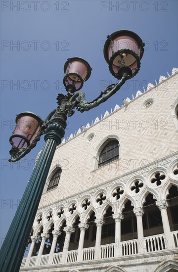 Venice, Veneto, Italy. Decorative street lamp in front of partly seen facade of the Palazzo Ducale against cloudless blue sky. Italy Italia Italian Venice Veneto Venezia Europe European City Architecture Facade Palazzo Ducale Palace Lamp Post Light Lights Streetlamp Blue sky Destination Destinations History Historic Southern Europe