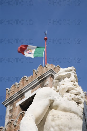 Venice, Veneto, Italy. Centro Storico Arsenale. Part view of crenellated tower flying Italian tricolour flag with statue of Neptune in foreground against clear blue sky of late summer. Italy Italia Italian Venice Veneto Venezia Europe European City Centro Storico Arsenale Tower Flag Tricolour Tricolor Statue Neptue Blue Sky Destination Destinations History Historic Southern Europe
