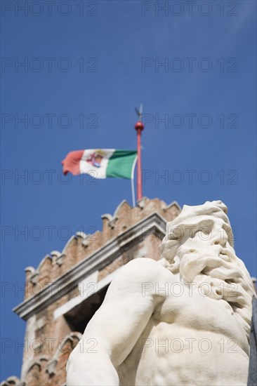 Venice, Veneto, Italy. Centro Storico Arsenale Part view of crenellated tower flying Italian tricolour flag with statue of Neptune in foreground against clear blue sky of late summer. Italy Italia Italian Venice Veneto Venezia Europe European City Sky Blue Neptue Statue Tricolor Tricolour Flag Tower Arsenale Storico Centro Destination Destinations History Historic Southern Europe