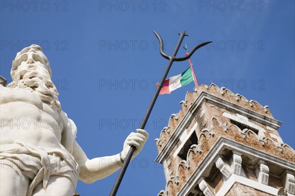 Venice, Veneto, Italy. Centro Storico Arsenale Part view of crenellated tower flying Italian tricolour flag with statue of Neptune in foreground against clear blue sky of late summer. Italy Italia Italian Venice Veneto Venezia Europe European City Centro Storico Arsenale Flag Tower Tricolour Tricolor Statue Neptue Blue Sky Destination Destinations History Historic Southern Europe
