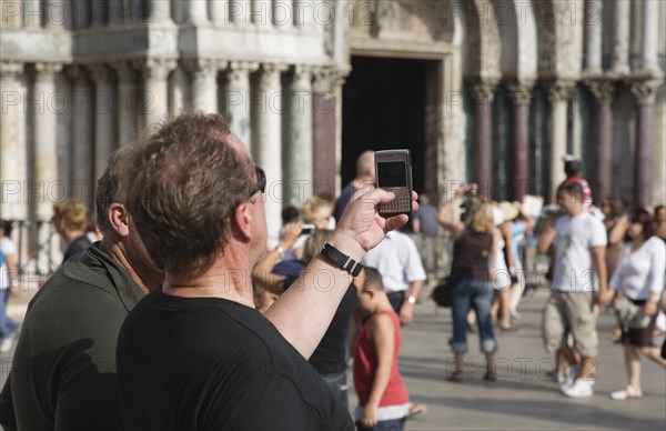 Venice, Veneto, Italy. Centro Storico St. Marks Square Tourist takes photograph on mobile phone with tourist crowds and facade of Basilica di San Marco part seen behind. Italy Italia Italian Venice Veneto Venezia Europe European City San Marco Basilico St Saint Mark Marks Square Tourist Tourists Mobile Phone Cell Phone Camera Pictures Snps Snaps Cellular Destination Destinations Holidaymakers Sightseeing Southern Europe Tourism