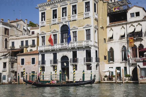 Venice, Veneto, Italy. Gondolier steers gondola from Rialto bridge along Grand Canal with Venice Republic EU and Italian tricolour flags on the facades of buildings behind. Italy Italia Italian Venice Veneto Venezia Europe European City Grand canal Water Gondola Gondolas Gondolier tourist Tourists Transport travel Architectures Facade Exterior Destination Destinations European Union Holidaymakers Sightseeing Southern Europe Tourism