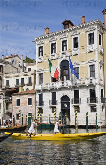 Venice, Veneto, Italy. Female pair taking part in Regata Storica Venice Regatta rowing yellow gondola on the Grand Canal passing waterside buildings with Venice Republic EU and Italian tricolour flags hanging from facades. Italy Italia Italian Venice Veneto Venezia Europe European City Regata Regatta Rowing Gondola Boat Architecture Facade Exterio Flag Flags Water Women Girls Female Blue Destination Destinations European Union Female Woman Girl Lady Southern Europe