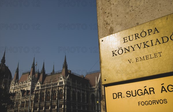 Budapest, Pest County, Hungary. Hungarian language signs on corner of office building in foreground with Parliament Building behind. Hungary Hungarian Europe European East Eastern Buda Pest Budapest City Urban Sign Builiding Parliament. Destination Destinations Eastern Europe Parliment Signs Display Posted Signage