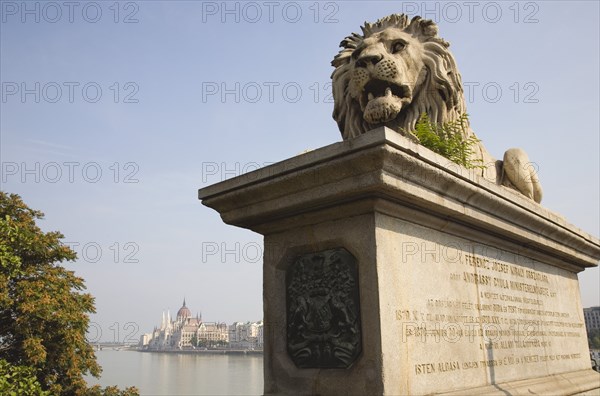 Budapest, Pest County, Hungary. Lion sculpture on the Chain Bridge with Hungarian Parliament Building on far Pest bank in background. Hungary Hungarian Europe European East Eastern Buda Pest Budapest City Transport Architecture Art Lion Statue Carving Sculpture Chain memory Bridge Parliament Building River Danube Destination Destinations Eastern Europe Parliment