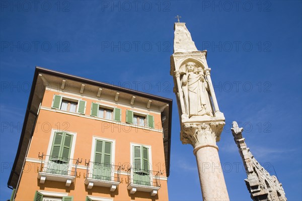 Verona, Veneto, Italy. Pastel painted house facade with pale green window shutters and balconies. Religious statue on free standing pillar in foreground against cloudless blue sky. Veneto Verona Europe European City Pastel Color Colour Colored Coloured Colorful Colourful Architecture Facade Exterior Window Windows Shuttered Shutters Statue Religious Religion Christian Blue Sky Destination Destinations Italia Italian Religion Religious Christianity Christians Southern Europe