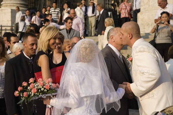 Budapest, Pest County, Hungary. Bride and groom greeting wedding guests on steps of Saint Stephens Basilica with groom leaning forward to kiss older man on cheek. Hungary Hungarian Europe European East Eastern Buda Pest Budapest City People Crowd Couple Wed Wedding Marriage Bride Groom White Dress Veil Flowers Color Cultural Cultures Eastern Europe Male Men Guy Marriage Marrying Espousing Hymeneals Nuptials Order Fellowship Guild Club Religion Religious