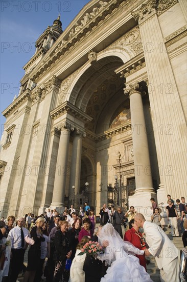 Budapest, Pest County, Hungary. Bride and groom greeting wedding guests on the steps of Saint Stephens Basilica after service both leaning forward to kiss older couple on cheek. Hungary Hungarian Europe European East Eastern Buda Pest Budapest City People Crowd Couple Wed Wedding Marriage Bride Groom White Dress Veil Flowers St Saint Stephen Stephens Basilica Church Exterior Blue Color Cultural Cultures Eastern Europe Marriage Marrying Espousing Hymeneals Nuptials Order Fellowship Guild Club Religion Religious