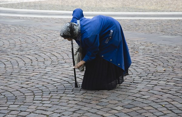 Budapest, Pest County, Hungary. Elderly woman bent over and supported with cane begging in square in front of Saint Stephen Basilica. Hungary Hungarian Europe European East Eastern Buda Pest Budapest City Woman Old Elderly Bent Crouched Over Down Walking Stick Beggar Begging Cobbled Street Road Cobbles Blue Grey Gray Eastern Europe Female Women Girl Lady Old Senior Aged One individual Solo Lone Solitary