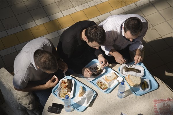 Budapest, Pest County, Hungary. Looking down on three young men seated at circular table having lunchtime meal of Hungarian sausages bread and pickles at the Central Market Nagy Vasarcsarnok. Mobile phones and bottled water visible on table. Hungary Hungarian Europe European East Eastern Buda Pest Budapest City Nagy Vasarcsarnok Central Market People Men Food Cafe Restaurant Eating Lunch Lunchtime Meal Table Seated Sat Tray Trays Sausage Sausages Bread Pickles Water Bottles Bottled 3 Bar Bistro Cell Cellular Eastern Europe Immature Male Man Guy