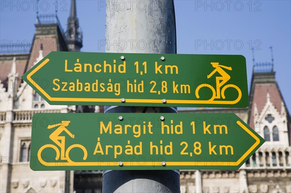Budapest, Pest County, Hungary. Green and yellow signs indicating direction of cycle lanes outside the neo-Gothic Parliament Building. Hungary Hungarian Europe European East Eastern Buda Pest Budapest City Architecture Building Parliament Sign Signs Green Yellow Cycle Lanes Cycling Bike Biking Bikes Cyclist Color Destination Destinations Eastern Europe Parliment Signs Display Posted Signage