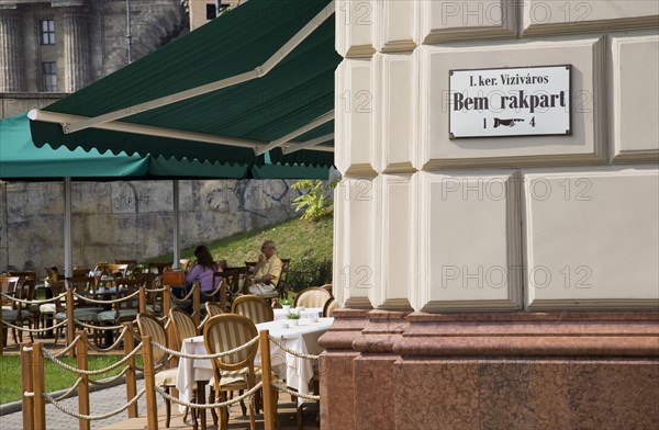 Budapest, Pest County, Hungary. Buda cafe beside the River Danube at approach to Chain Bridge with people seated at outside tables. Hungary Hungarian Europe European East Eastern Buda Pest Budapest City Cafe People Sat Outside Exterior Tables Bar Bistro Destination Destinations Eastern Europe Restaurant