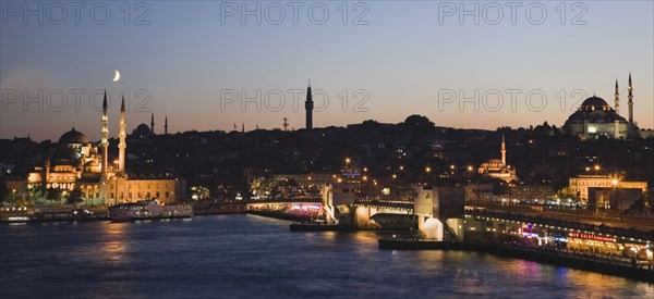 Istanbul, Turkey. Sultanahmet. The Golden Horn. The New Mosque or Yeni Camii at left the Galata Bridge and Suleymaniye Mosque at right with buildings and restaurants illuminated at dusk and crescent moon in blue purple sky above. Turkey Turkish Istanbul Constantinople Stamboul Stambul City Europe European Asia Asian East West Urban Skyline Destination Travel Tourism Sultanahmet Golden Horn New Mosque Yeni Camii Galata Bridge Suleymaniye Dusk Sunset Night Illuminated Skyline Minarets Water Crescent Moon Citiscape Building Buildings Urban Architecture Destination Destinations Middle East Nightfall Twilight Evenfall Crespuscle Crespuscule Gloam Gloaming Nite Religious South Eastern Europe Sundown Atmospheric Turkiye Western Asia