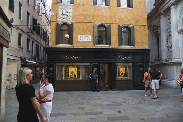 Venice, Veneto, Italy. Centro Storico Cartier jewellery store exterior with security guard and tourists in paved square with shuttered facade and plaque depicting St George and the dragon. Italy Italia Italian Venice Veneto Venezia Europe European City Centro Storico Cartier Jewellery Jewllery Store Shop Campo S.Zulian Square Tourist Tourists People Security Guard Guards Destination Destinations Holidaymakers Jewelry Sightseeing Southeast Asia Southern Europe Tourism