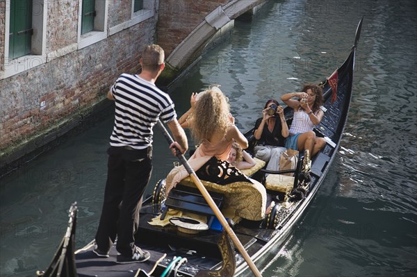 Venice, Veneto, Italy. Centro Storico Gondolier steering gondola along canal as models are photographed for fashion shot in late summer sunshine. Italy Italia Italian Venice Veneto Venezia Europe European City Centro Storico Gondola Gondolas Gondolier Tourist Tourists Women Taking Photographs Pictures Snap Snaps Portraits Holiday Vacation Canal Water Destination Destinations Female Woman Girl Lady Holidaymakers Sightseeing Southern Europe Tourism
