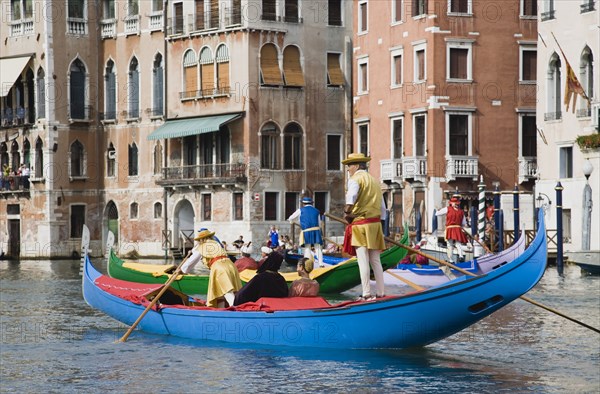 Venice, Veneto, Italy. Participants in the Regata Storico annual historical regatta in brightly painted gondolas and wearing traditional costume approaching the Rialto bridge with onlookers on balconies of canalside buildings behind. Teams represent Sestiere districts of Venice in traditional races. Italy Italia Italian Venice Veneto Venezia Europe European City Regata Regatta Gondola Gondola Gondolas Gondolier Boat Architecture Exterior Water Classic Classical Color Destination Destinations History Historic Older Southern Europe