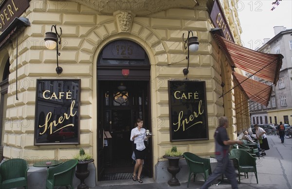 Vienna, Austria. Mariahilf District. Cafe Sperl the preferred cafe of Adolf Hitler. Exterior facade with waitress carrying coffee order at entrance. Striped awning pulled out at side with customers sitting at outside tables on street below. Austria Austrian Vienna Viennese Wien Mariahlf 6th District Cafe Sperl Hitler Waitress Entrance Coffee Exterior Europe European Work Urban City Table Tables Schanigarten Bar Bistro Destination Destinations History Historic Osterreich Restaurant Viena Western Europe