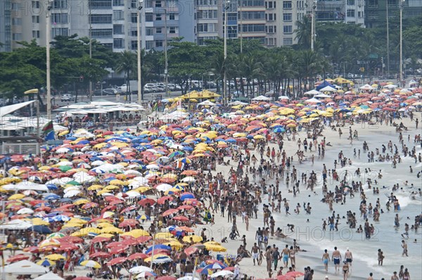 Rio de Janeiro, Brazil. Copacabana beach. Crowds on the beach and in the sea and multi-coloured beach umbrellas with part seen apartment buildings behind. Brazil Brasil Brazilian Brasilian South America Latin Latino American City Travel Destination Urban Vacation Beach Beaches People Sunbathing Crowds Rio de Janeiro Copacabana Umbrellas Parasols Destination Destinations Flat Holidaymakers Latin America Sand Sandy Beach Tourism Seaside Shore Tourist Tourists Vacation Sand Sandy Beaches Tourism Seaside Shore Tourist Tourists Vacation South America Southern Sunbather Water