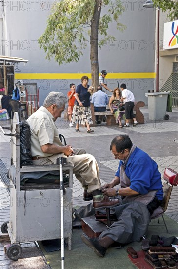 Santiago, Chile. Shoe-shine in downtown shopping area. Partially sighted or blind elderly man seated in chair with white stick at side to have his shoes cleaned brushes at side. Chile Chilean South America American Hispanic Santiago City Cityscape Urban Street Polish Shoes Shoe Shining Shine Cleaning Clean Working Work Men Man Cleansing Destination Destinations Latin America Latino Male Man Guy Male Men Guy Old Senior Aged Shops Shoppers Mall Retail Buy Buying Market Markets South America Southern Washing