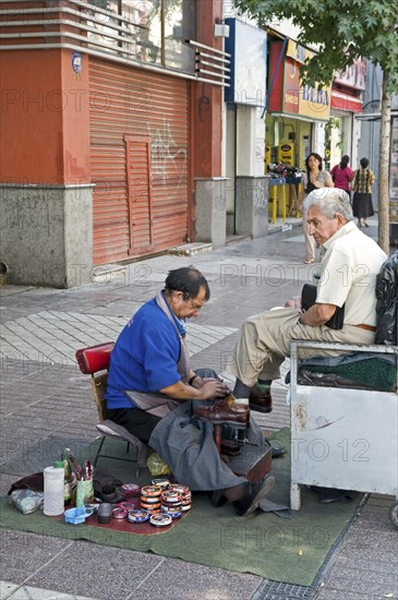 Santiago, Chile. Elderly man at street shoe shine in downtown shopping area piles of shoe polish tins at side. Chilean South America American Hispanic Santiago City Cityscape Urban Street Polish Shoes Shoe Shining Shine Cleaning Clean Working Work Men Man Cleansing Destination Destinations Latin America Latino Male Man Guy Male Men Guy Old Senior Aged Shops Shoppers Mall Retail Buy Buying Market Markets South America Southern Washing