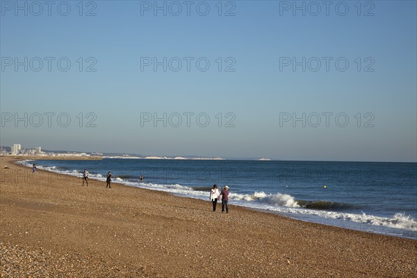 Shoreham-by-Sea, West Sussex, England. People walking along the waters edge on Shoreham beach. England English UK United Kingdom GB Great Britain British Europe European West Sussex County Shoreham Shoreham-by-Sea By Sea Beach People Couple Couples Walk Walking Stroll Strolling Shore Pebble Pebbles Blue Sky