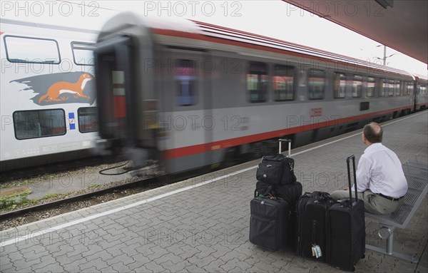 Vienna, Austria. Wien Westbahnhof. Passenger with luggage sitting on Vienna West Station with passing train. Also known as Vienna Western Station this is a major Austrian station and the starting point of the Austrian Western Railway or Westbahn for trains to Salzburg Munich Frankfurt Zurich Budapest Bucharest Belgrad etc. Austria Austrian Republic Vienna Viennese Wien Europe European City Capital Westbahnhof Transport Rail Railway Train Station Platform Commuter Tourist Luggage Baggage Bags man Destination Destinations Male Men Guy MŸnchen One individual Solo Lone Solitary Osterreich Sightseeing Tourists Viena Western Europe