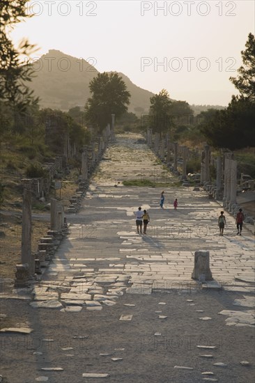 Selcuk, Izmir Province, Turkey. Ephesus. Tourists on paved road lined by broken columns in late afternoon sun in antique city of Ephesus on the Aegean sea coast. Turkey Turkish Eurasia Eurasian Europe Asia Turkiye Izmir Province Selcuk Ephesus Ruin Ruins Roman Column Columns Facde Ancient Architecture Masonry Rock Stone Paved Road Street Tourists Sunset Destination Destinations European History Historic Holidaymakers Middle East South Eastern Europe Sundown Atmospheric Tourism Water Western Asia
