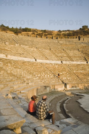 Selcuk, Izmir Province, Turkey. Ephesus. Tourist couple sitting in amphitheatre in the antique city of Ephesus on the Aegean sea coast. Turkey Turkish Eurasia Eurasian Europe Asia Turkiye Izmir Province Selcuk Ephesus Ruin Ruins Roman Amphitheatre Aamphitheater Ancient Architecture Masonry Rock Stone Tourists Destination Destinations European History Historic Holidaymakers Middle East Sightseeing South Eastern Europe Tourism Water Western Asia