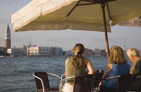 Venice, Veneto, Italy. Giudecca island Tourists enjoying lunch at terrace cafe in Centro Storico with view to St. Marks from shade of sun umbrella in late summer. Italy Italia Italian Venice Veneto Venezia Europe European City Water Giudecca Island Cafe Terrace Centro Stroico St Saint Mark Marks Umbrella Parasol People Tourists Bar Bistro Destination Destinations Holidaymakers Restaurant Southern Europe Tourism