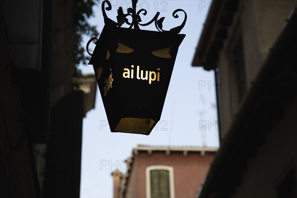 Venice, Veneto, Italy. Centro Storico Lantern outside restaurant inscribed with ai lupi which translates as to the wolves with reference to Dantes use of wolves as metaphor in the Inferno. Italy Italia Italian Venice Veneto Venezia Europe European City Centro Storico Latern Ai Lupi Wolves Restaurant Sign Illuminated Destination Destinations History Historic Signs Display Posted Signage Southern Europe