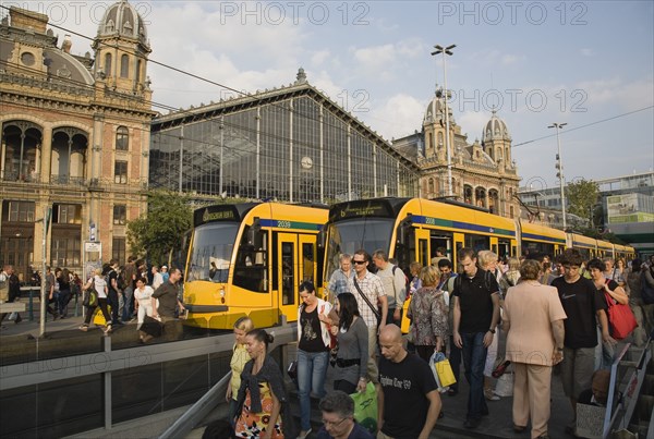 Budapest, Pest County, Hungary. Trams and crowd of passengers in front of rail terminus Budapest Nyugati palyaudvar and walking down steps to metro in foreground. Man appears to be begging on right hand side of steps unacknowledged by passers-by. Hungary Hungarian Europe European East Eastern Buda Pest Budapest City Transport Rail Train Tram Station Terminus Stop Nyugati Palyaudvar People Crowd Crowded Commuters Tourists Travel Passengers Descend Metro Underground Destination Destinations Eastern Europe Holidaymakers Male Men Guy Roundel Tourism